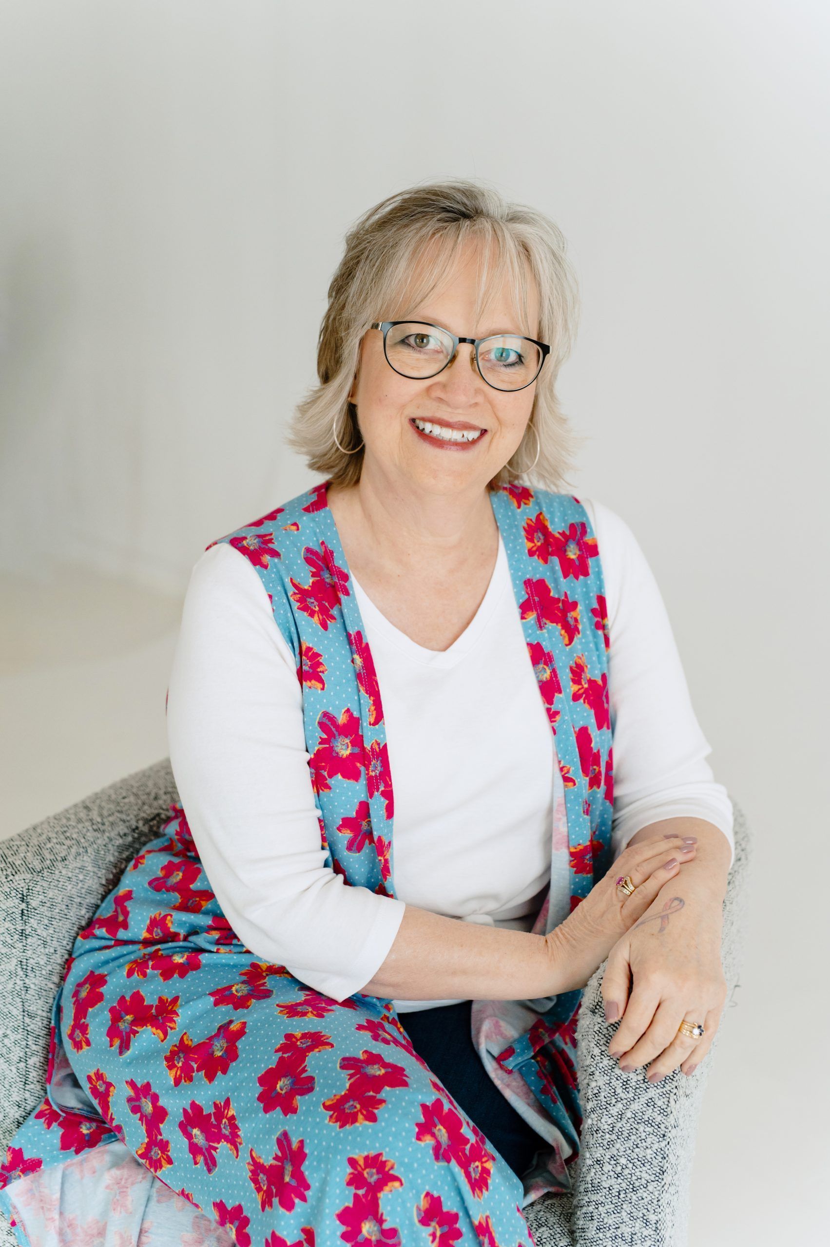 Smiling woman in glasses seated in a gray chair, wearing a white shirt and pink floral vest.