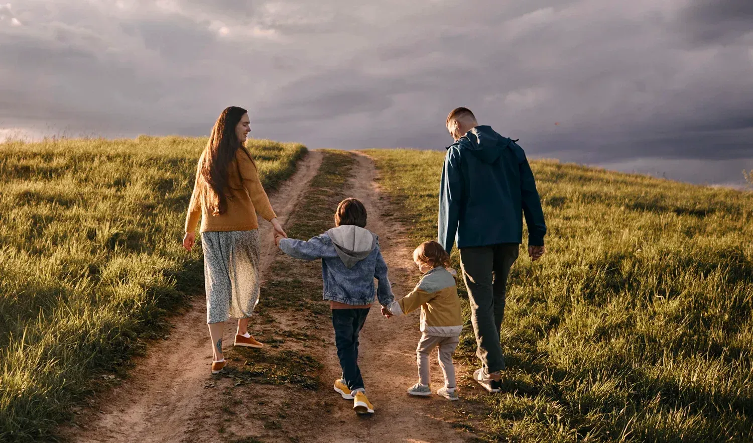 A family of four walking down a dirt path through grassy hills under a cloudy sky.