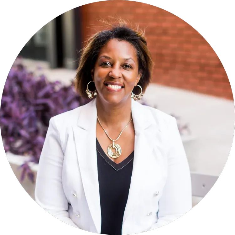 Smiling woman in a white blazer standing outdoors by a brick wall and purple flowers
