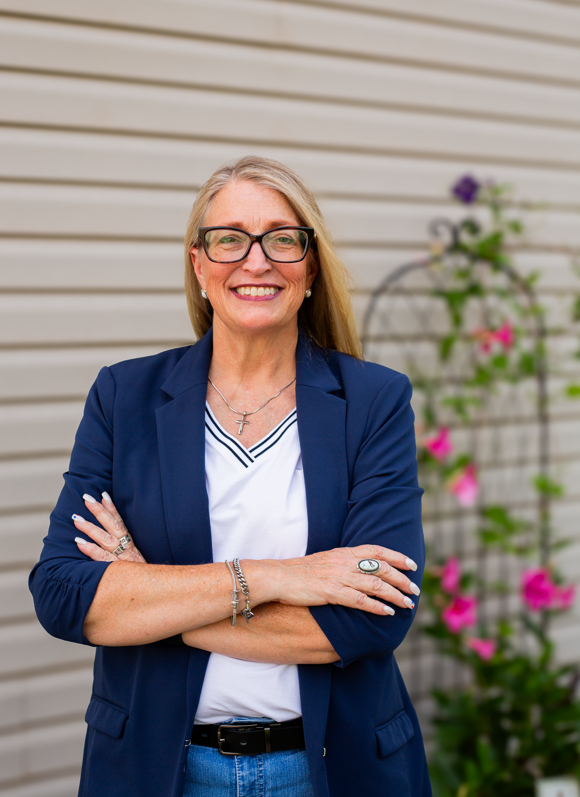 Smiling woman in a blue blazer standing with arms crossed against a light siding wall with flowers nearby