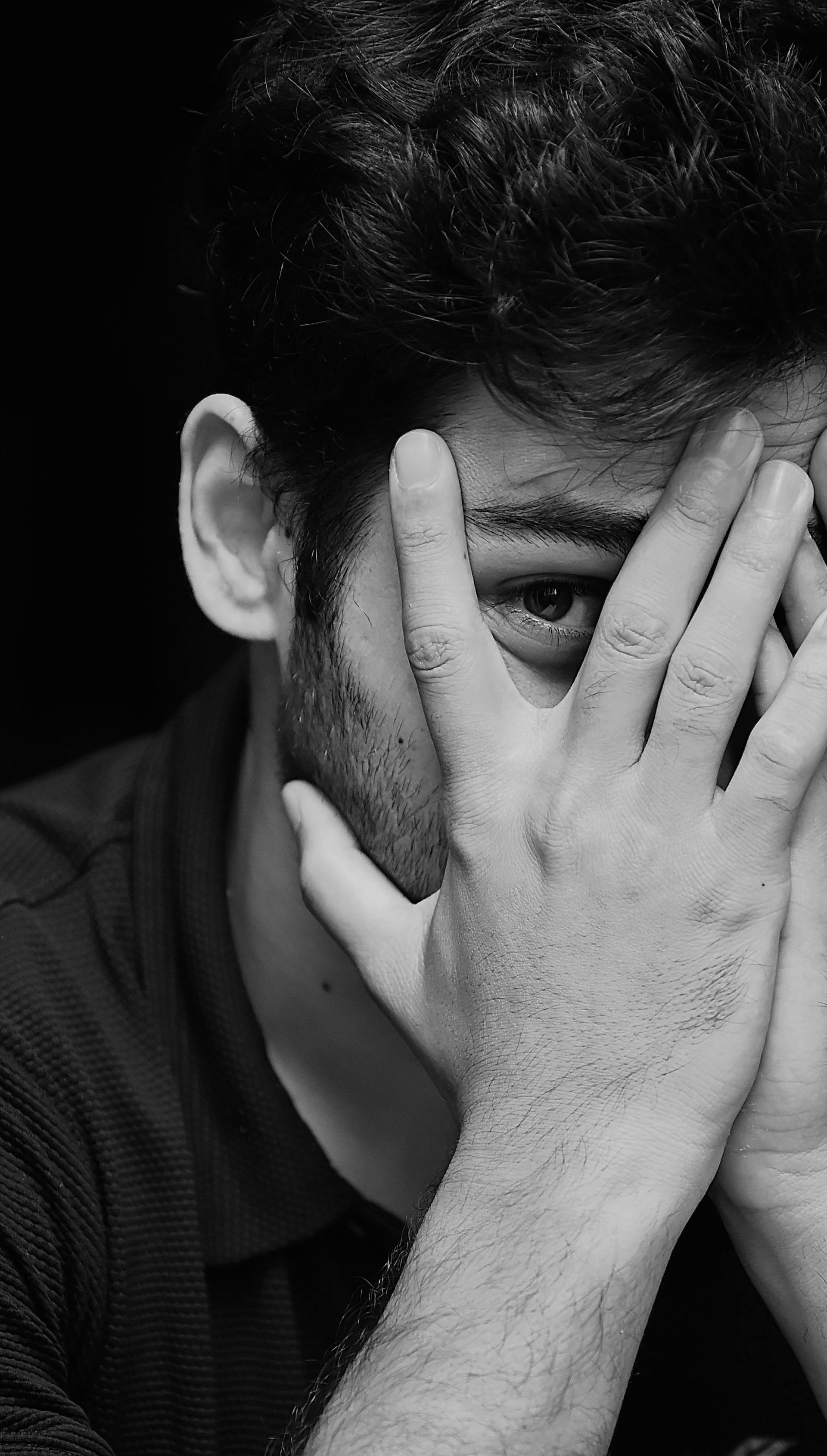 Black-and-white portrait of a person covering their face with both hands, partially revealing one eye