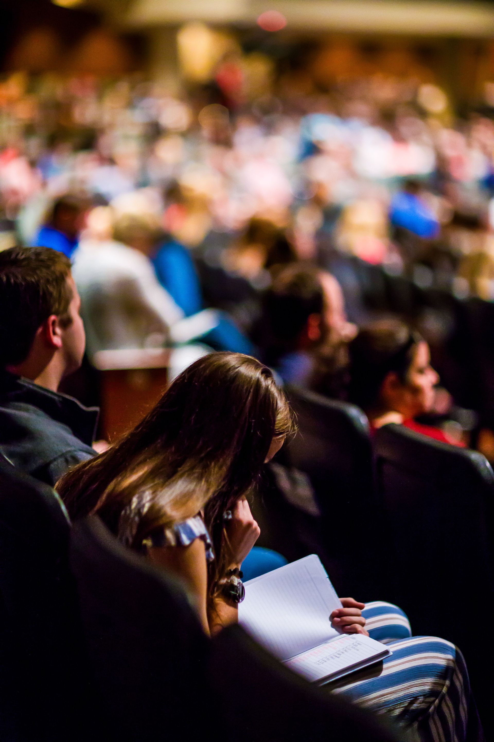 Audience seated in a dim auditorium, one person reading a program while others watch ahead