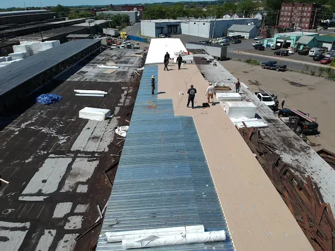 Aerial view of workers on a long rooftop under construction with blue and tan roofing materials.