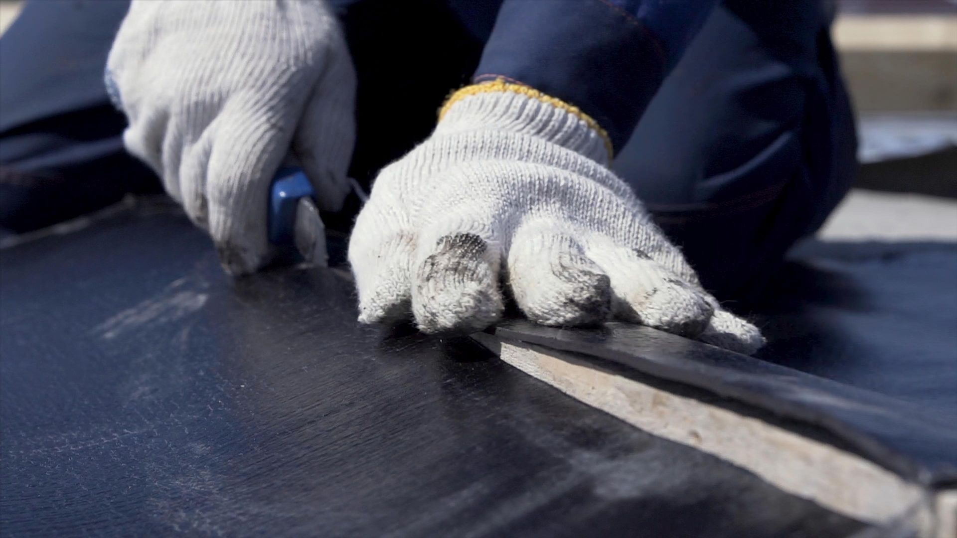 Gloved hands using a utility knife to cut black roofing material
