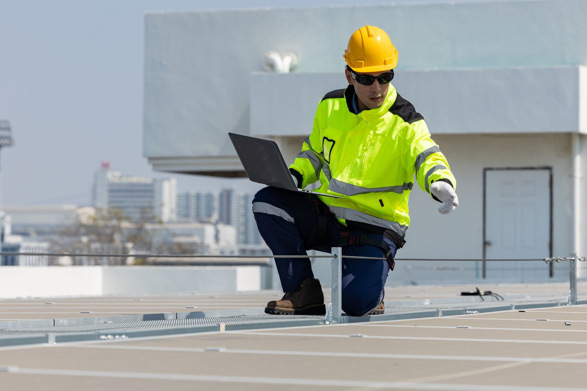 Worker in yellow hard hat inspecting rooftop with a laptop and tool on a sunny day