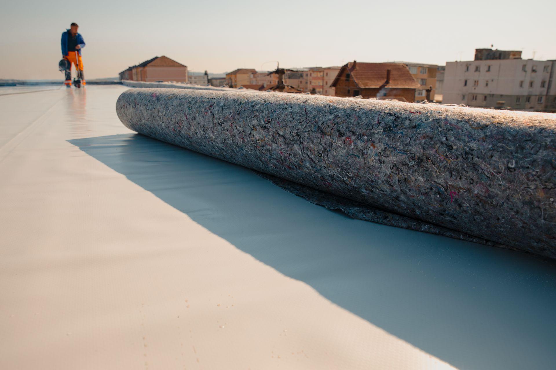 Person standing on a flat rooftop beside a large rolled mat, with buildings in the background.
