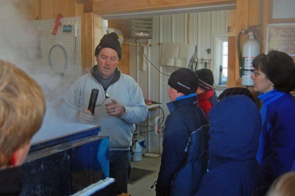Man demonstrating maple syrup making to a group. Steam rises from a blue container. Inside, rustic setting.