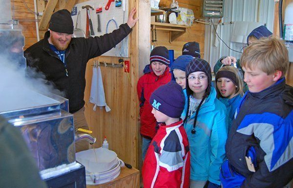 A man points to maple syrup boiling, explaining it to a group of children in a sugar shack.