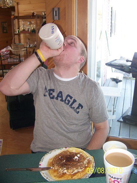 Man drinking from a container, eating pancakes. He wears a grey shirt, sitting at a table near a window.