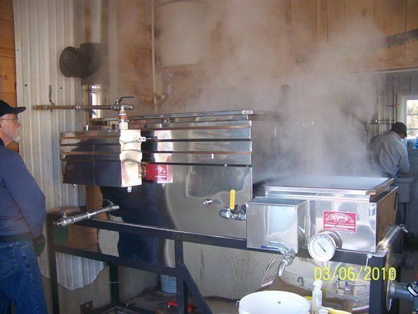 Maple syrup evaporator steaming, man watches. Stainless steel equipment, shed setting.