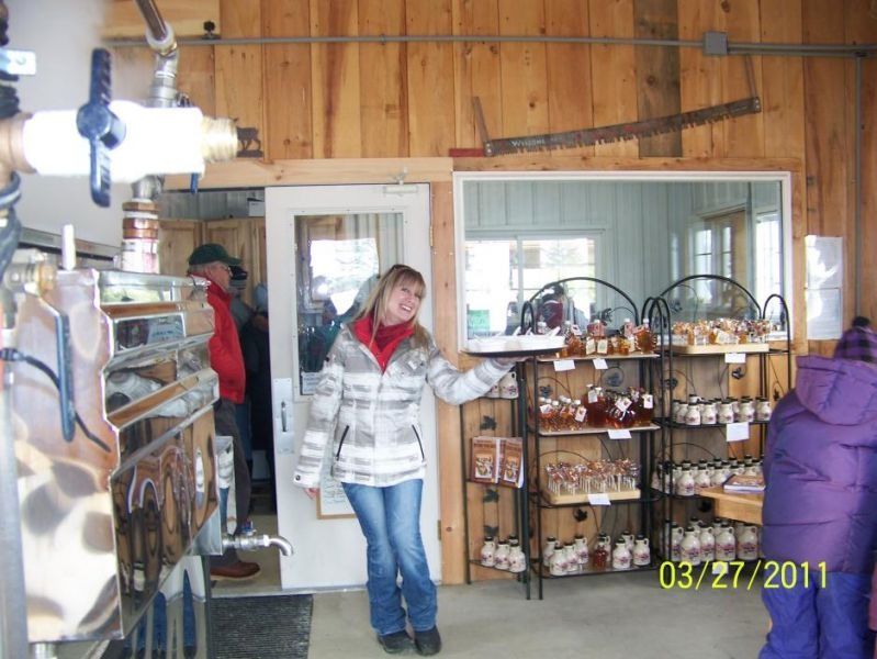 Woman holding tray of maple syrup products in a shop; products on shelves, man in doorway.