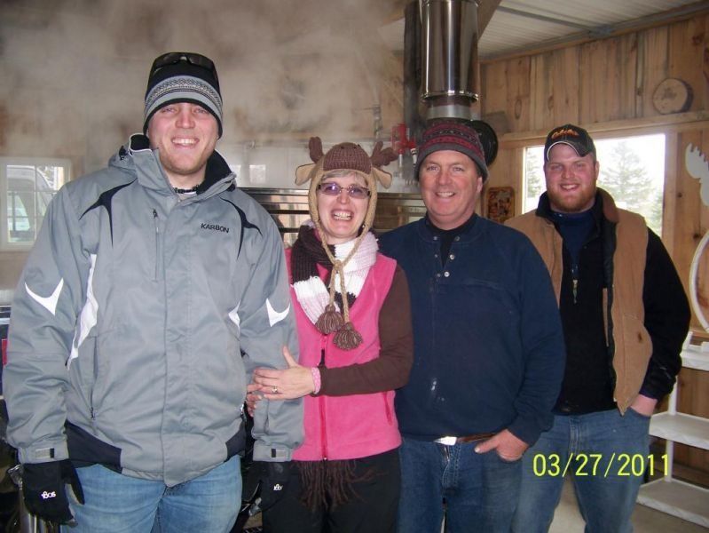Four people smiling in front of maple syrup evaporator, all wearing winter hats.