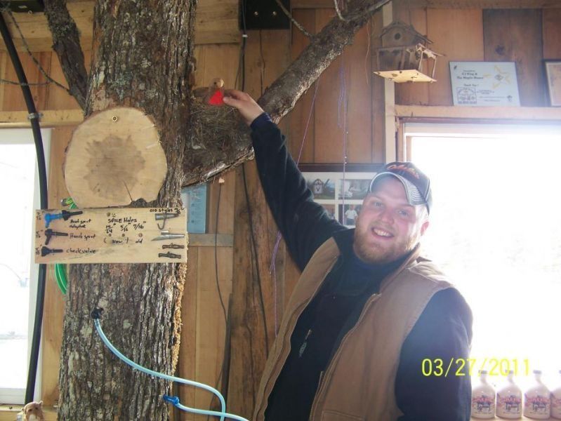Man in a treehouse, reaching towards a red object. He smiles near a tree, tap, and sign.
