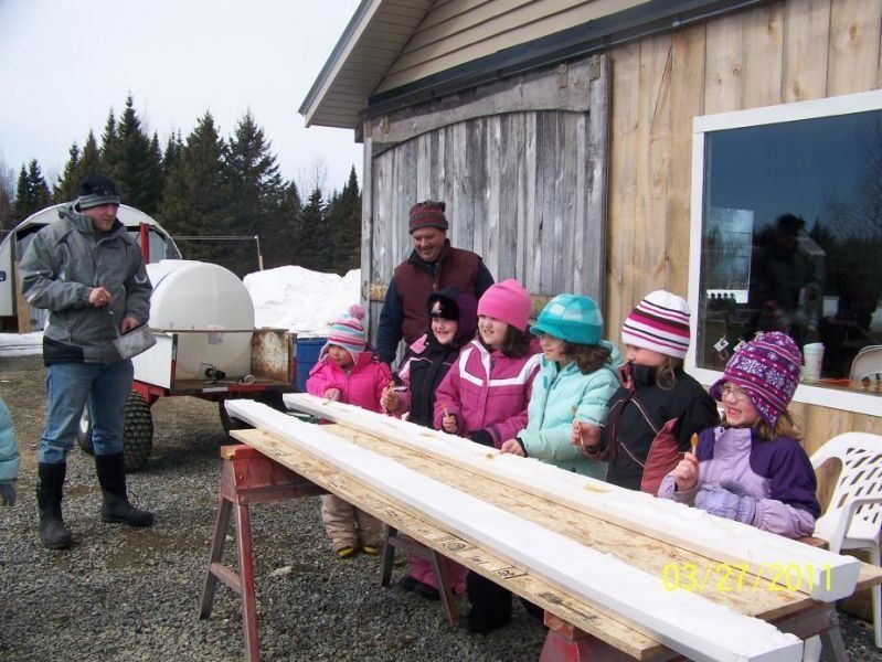 Children and adults stand near wood planks outdoors. They wear winter hats and coats.