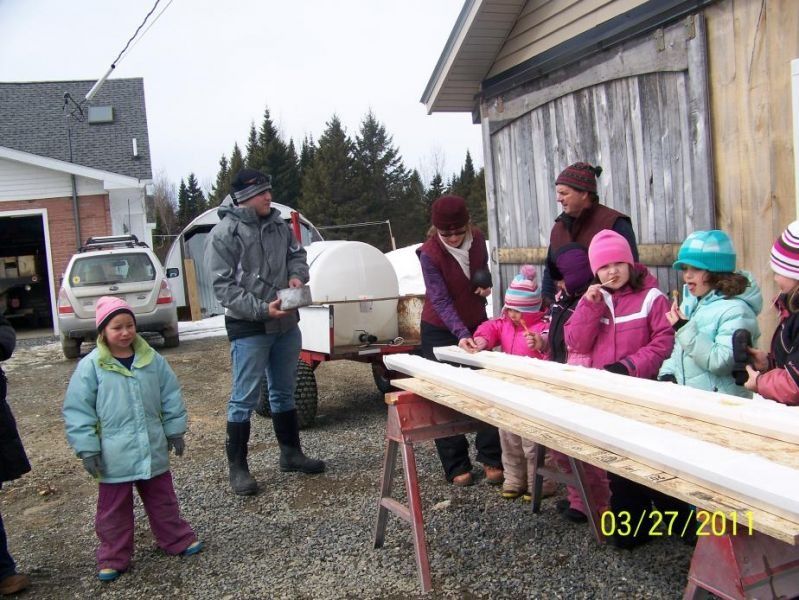 Children and adults outdoors, looking at lumber on sawhorses. White building and car in the background.