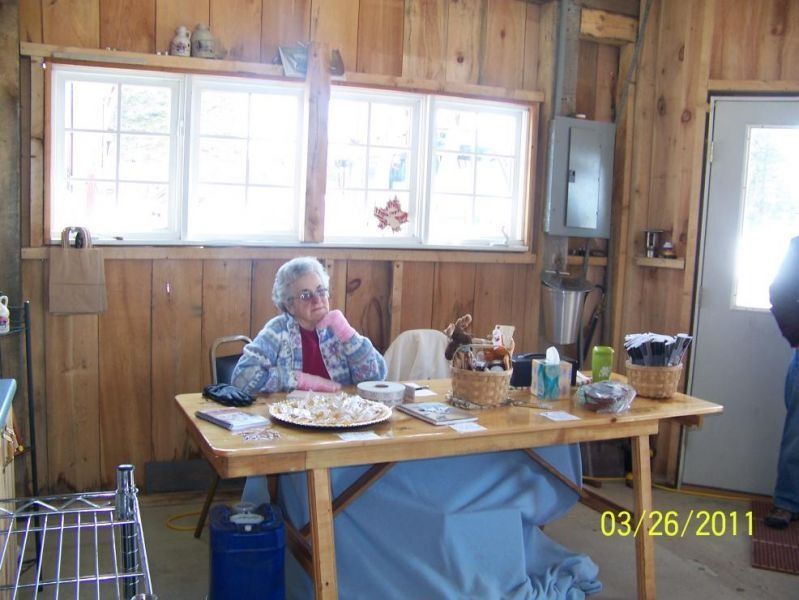 An elderly person sits at a wooden table in a rustic room. They rest their chin on their hand near treats.