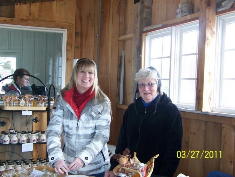 Two women smiling in a maple syrup shop, beside a display of products. Interior setting with wooden walls.
