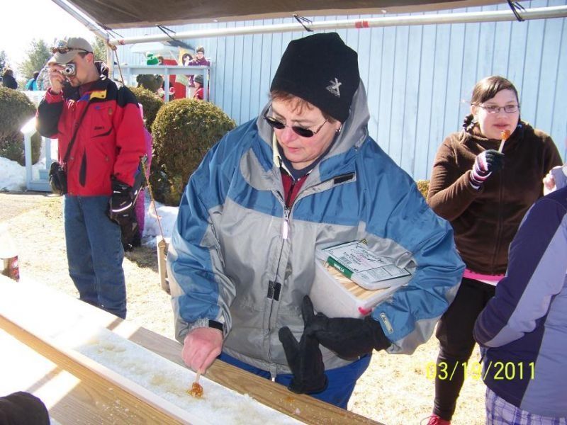 Person in blue jacket adds maple syrup to snow at outdoor event. Others watch and enjoy the sweet treat.