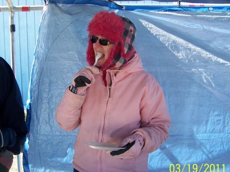 Woman in pink winter wear eating with a fork; blue tarp backdrop.