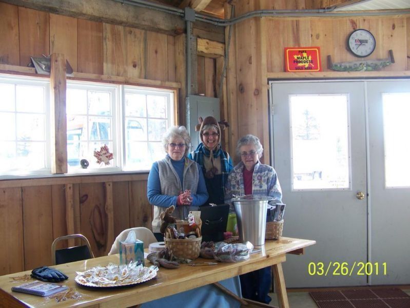 Three women stand behind a table with items, inside a wooden-walled building.