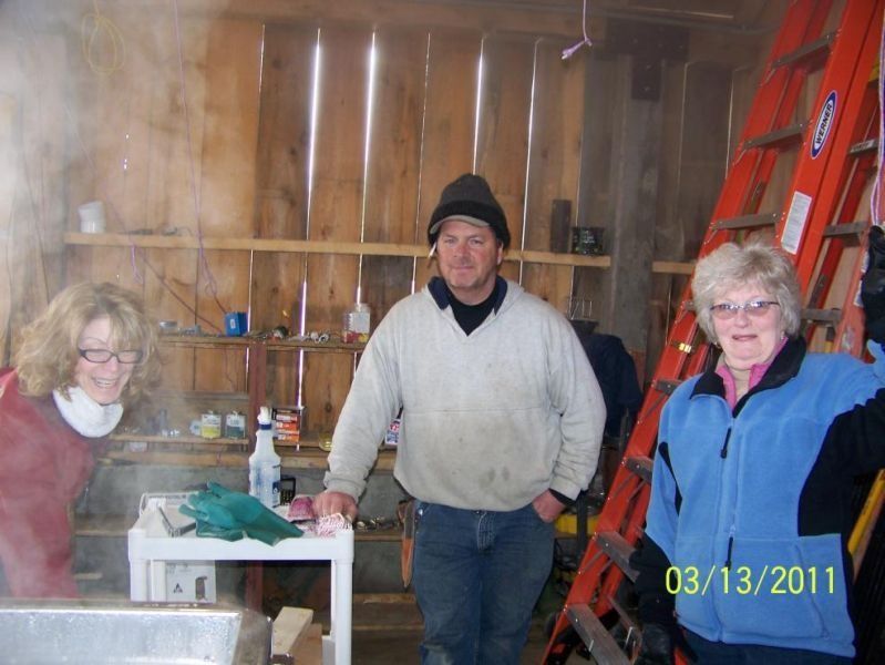 Three people in a sugar shack. One man and two women, smiling near steaming equipment, next to an orange ladder.