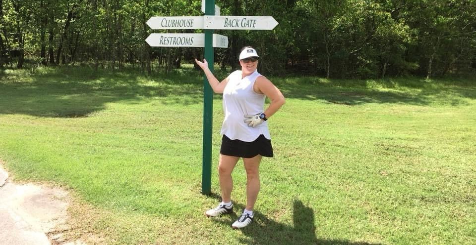 A woman is standing next to a sign in a grassy field.