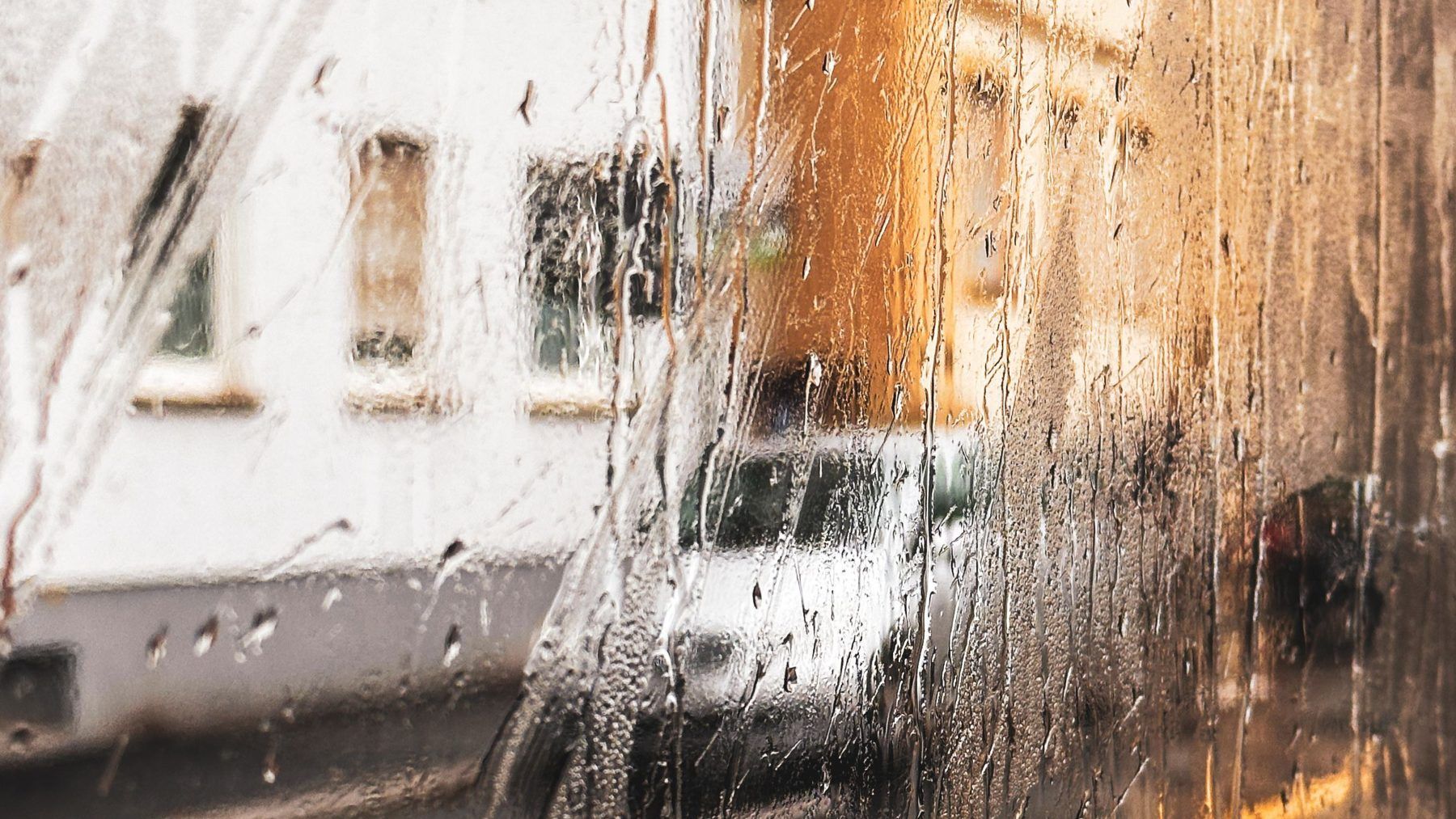 Rain running down a car window with parked cars and buildings beyond
