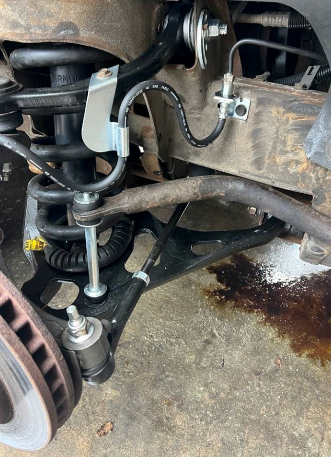 Close-up of a car’s front suspension, brake rotor, and control arm with grease on the ground. | Mike's Garage