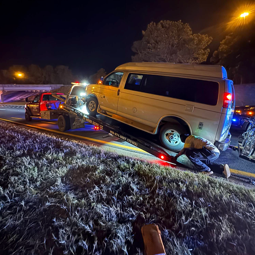 Tow truck loading a white van on a highway at night, with a worker crouched beside it and flashing lights nearby