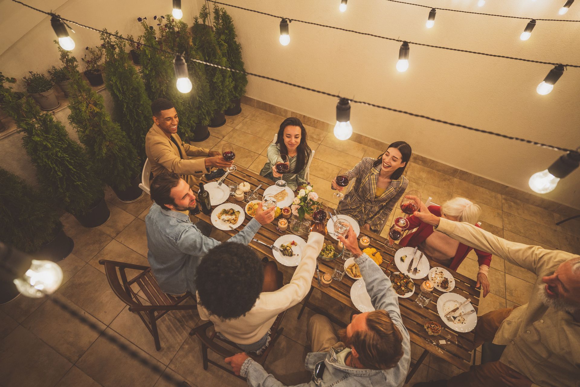 Des amis partagent un dîner aux chandelles à une table extérieure éclairée par des guirlandes lumineuses, avec de la nourriture et des boissons.