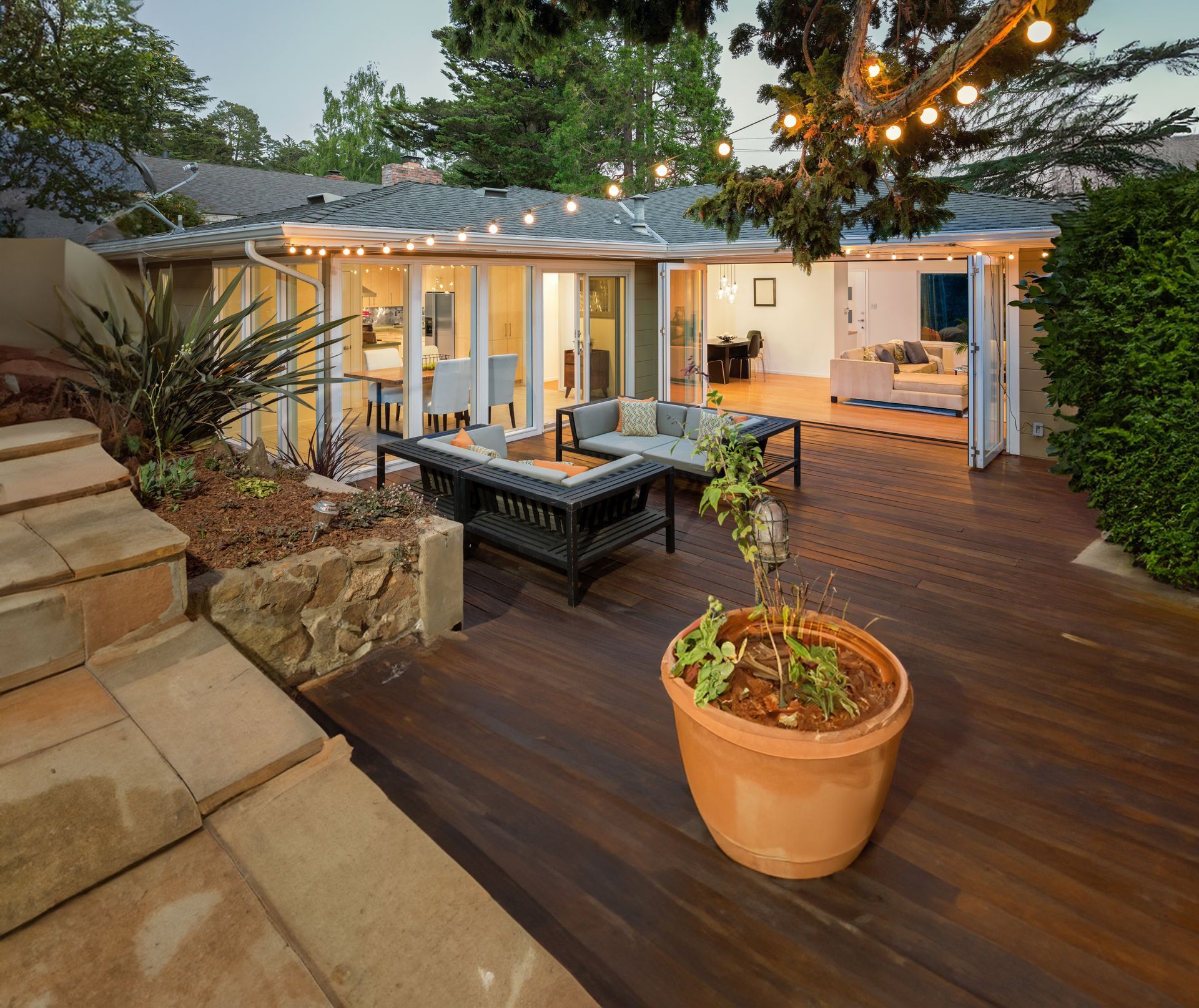 Terrasse en bois dans le jardin avec sièges extérieurs, plantes en pot et guirlandes lumineuses à côté d'une maison moderne