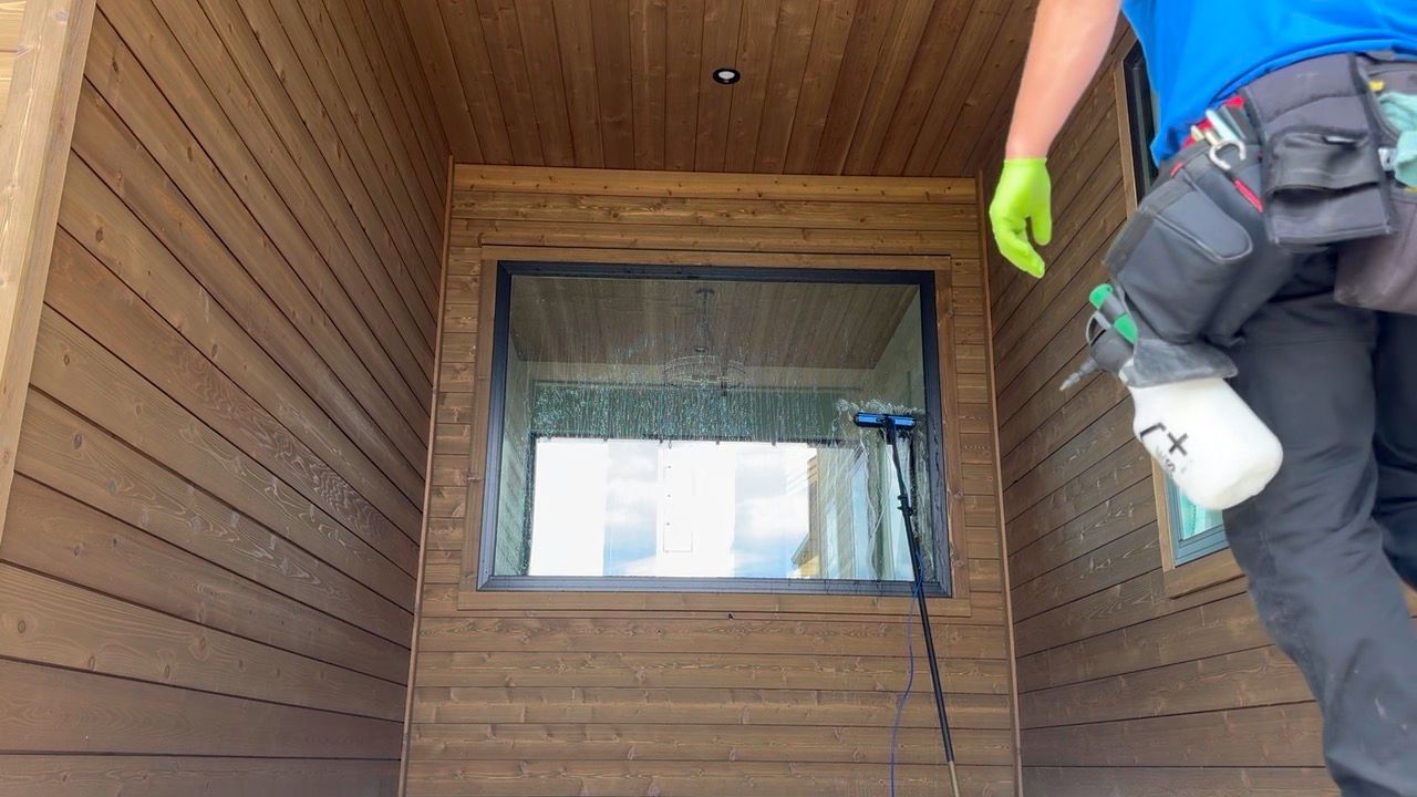 Worker with green gloves and spray tank at a wooden porch window, cleaning or painting the trim