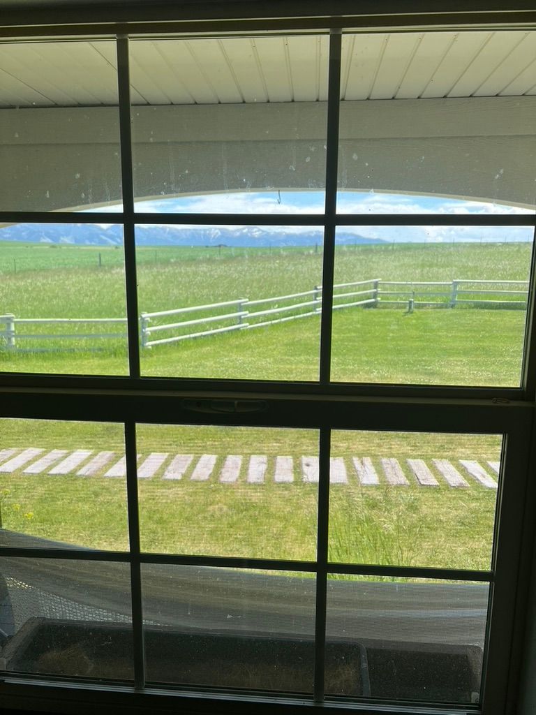 View through a window of a green lawn, white fence, and distant water under a cloudy sky