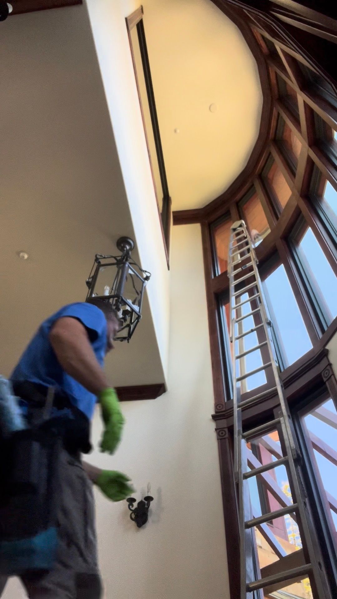Worker in blue shirt and green gloves repairing a ceiling in a tall stairwell with large windows.