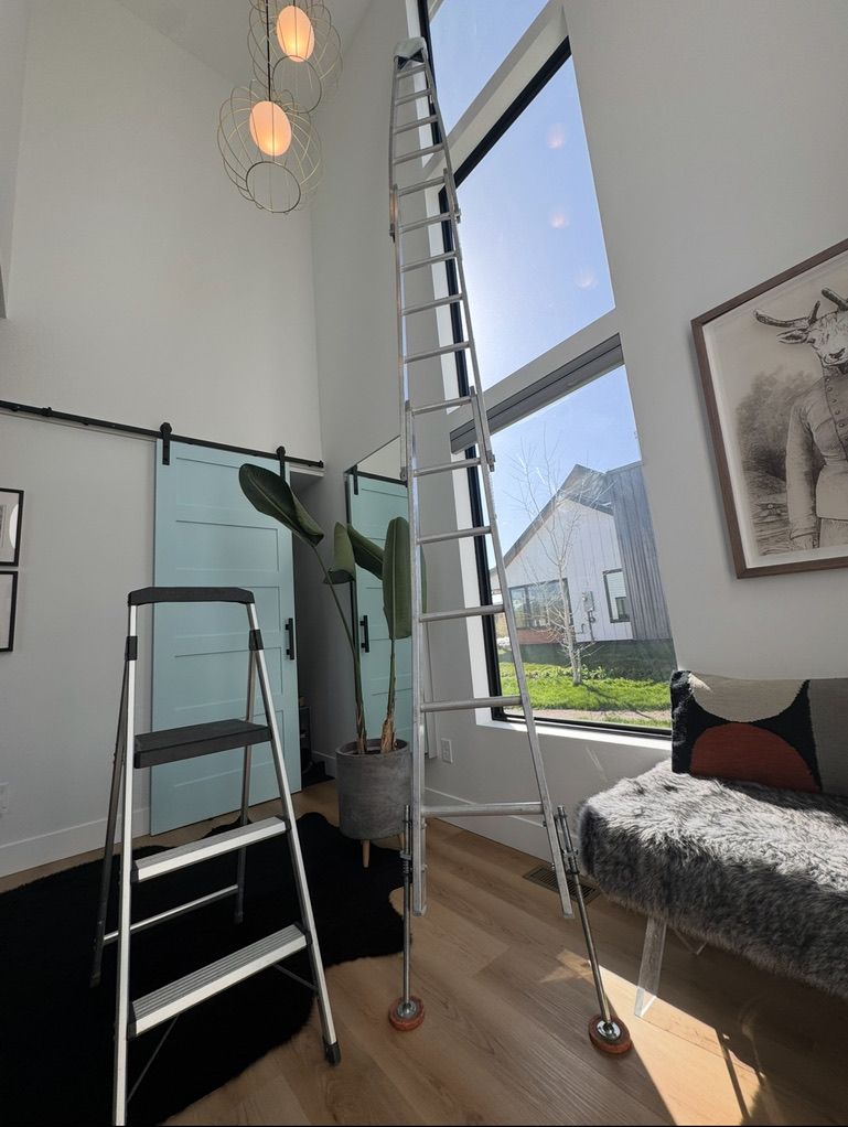 Loft bedroom with ladder, tall window, bed, and glass partition, lit by bright daylight