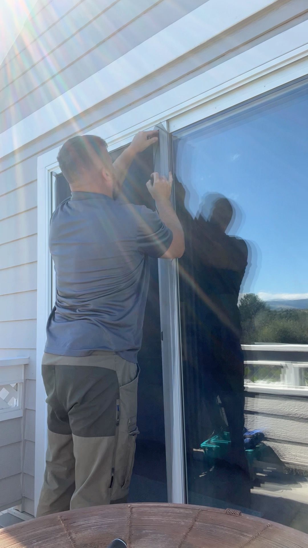 Two people installing or adjusting a window screen on the side of a house in bright sunlight