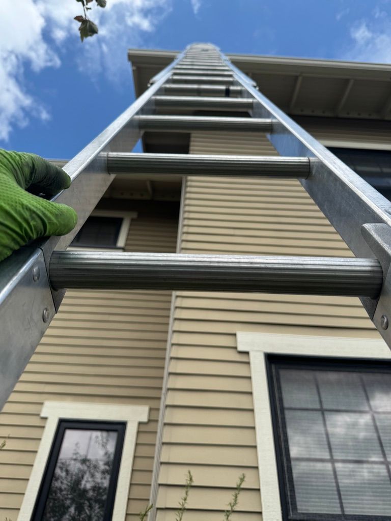 Person on a ladder cleaning a house exterior under a blue sky