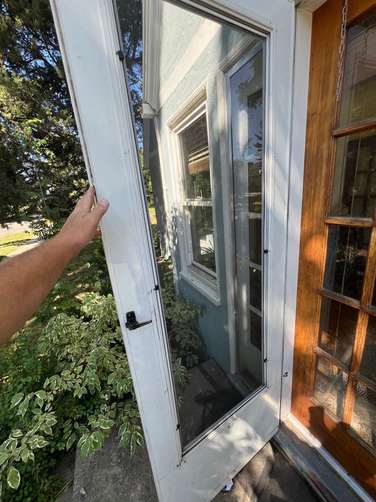 Hand holding open a white screen door beside a house window and brick siding