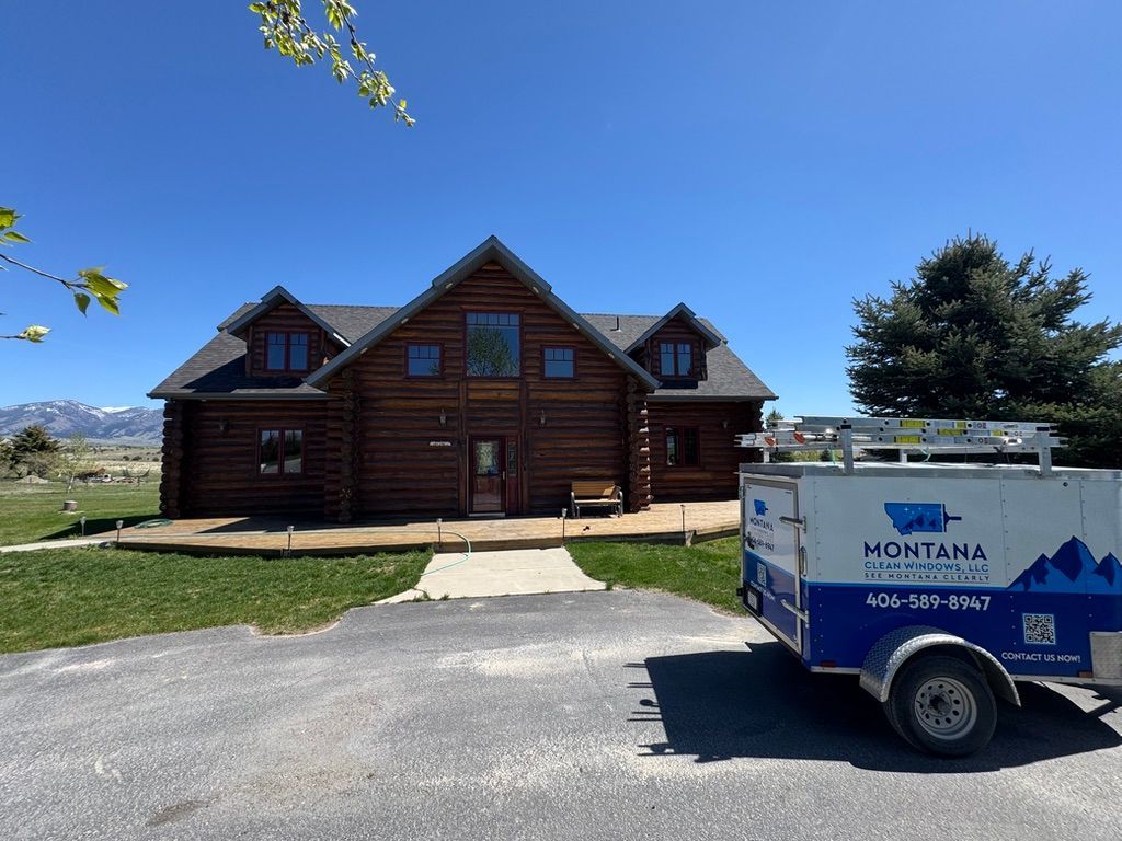 Log cabin house with a Montana utility trailer parked on a gravel driveway under a clear blue sky