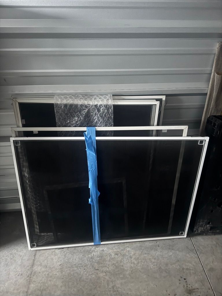 Stacked black-framed window screens leaning against a gray wall, secured with a blue strap.
