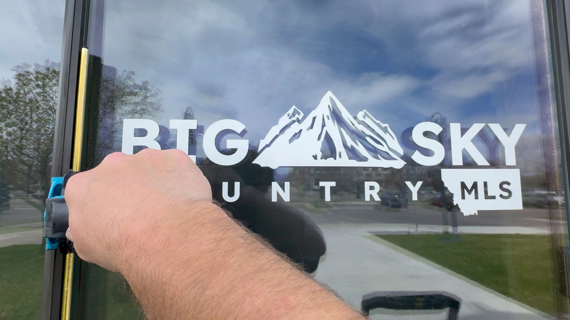 Hand holding a black marker on a vehicle window with the Big Sky Country MLS logo decal.
