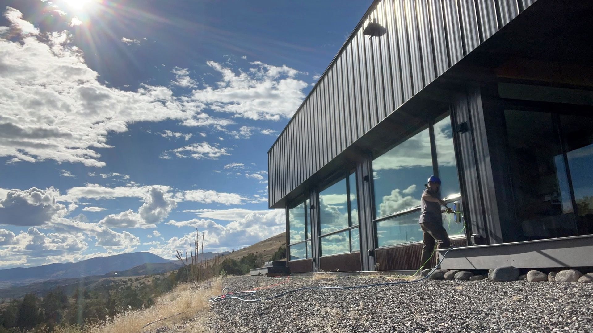 Modern glass building on a sunny hillside with mountains and a person standing outside.