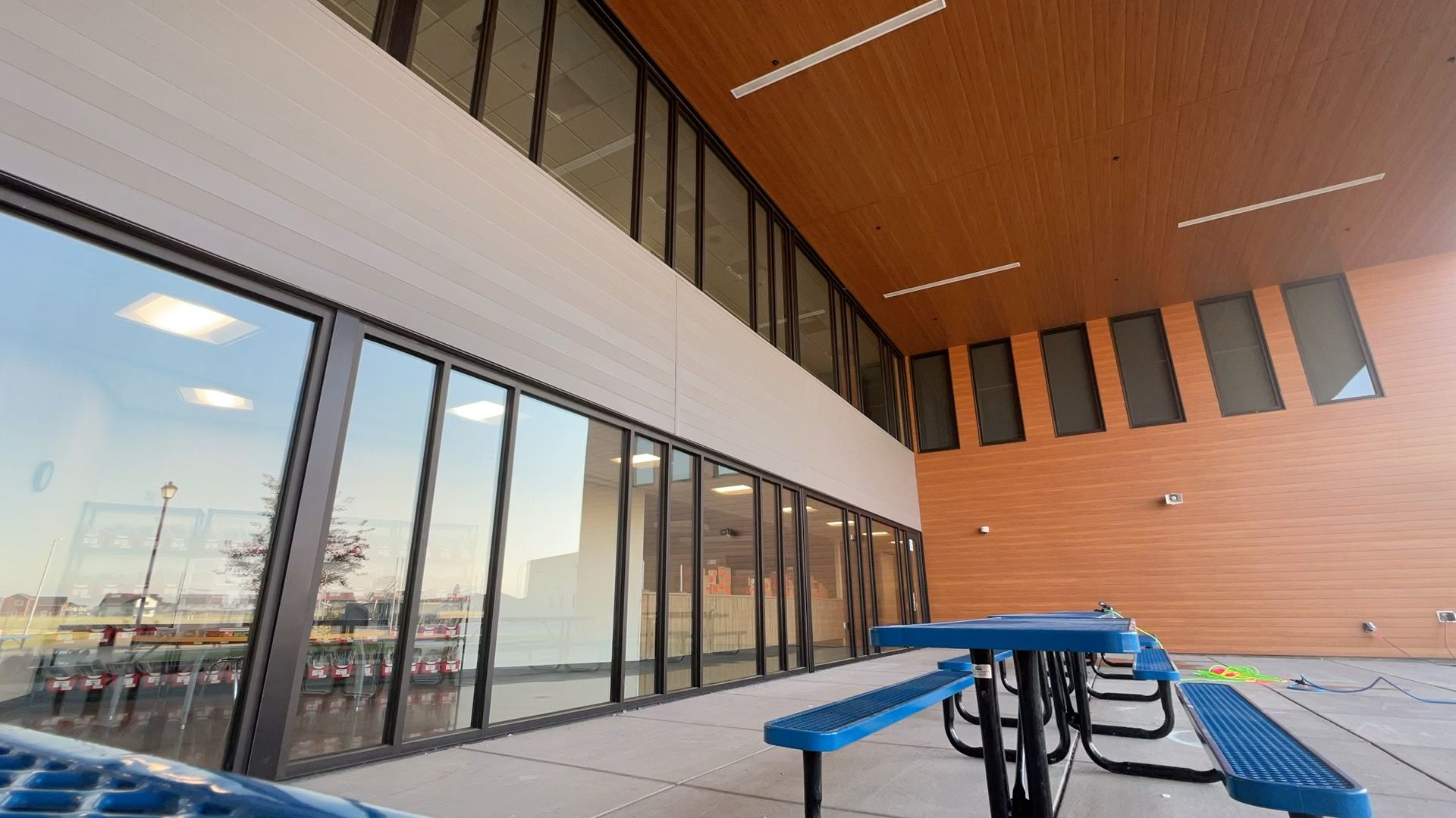 Covered outdoor patio with blue picnic tables beside large glass windows and a wood-paneled wall