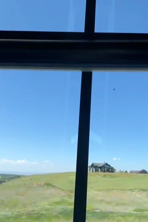 View through a window of a green hillside under a clear blue sky, with a house in the distance