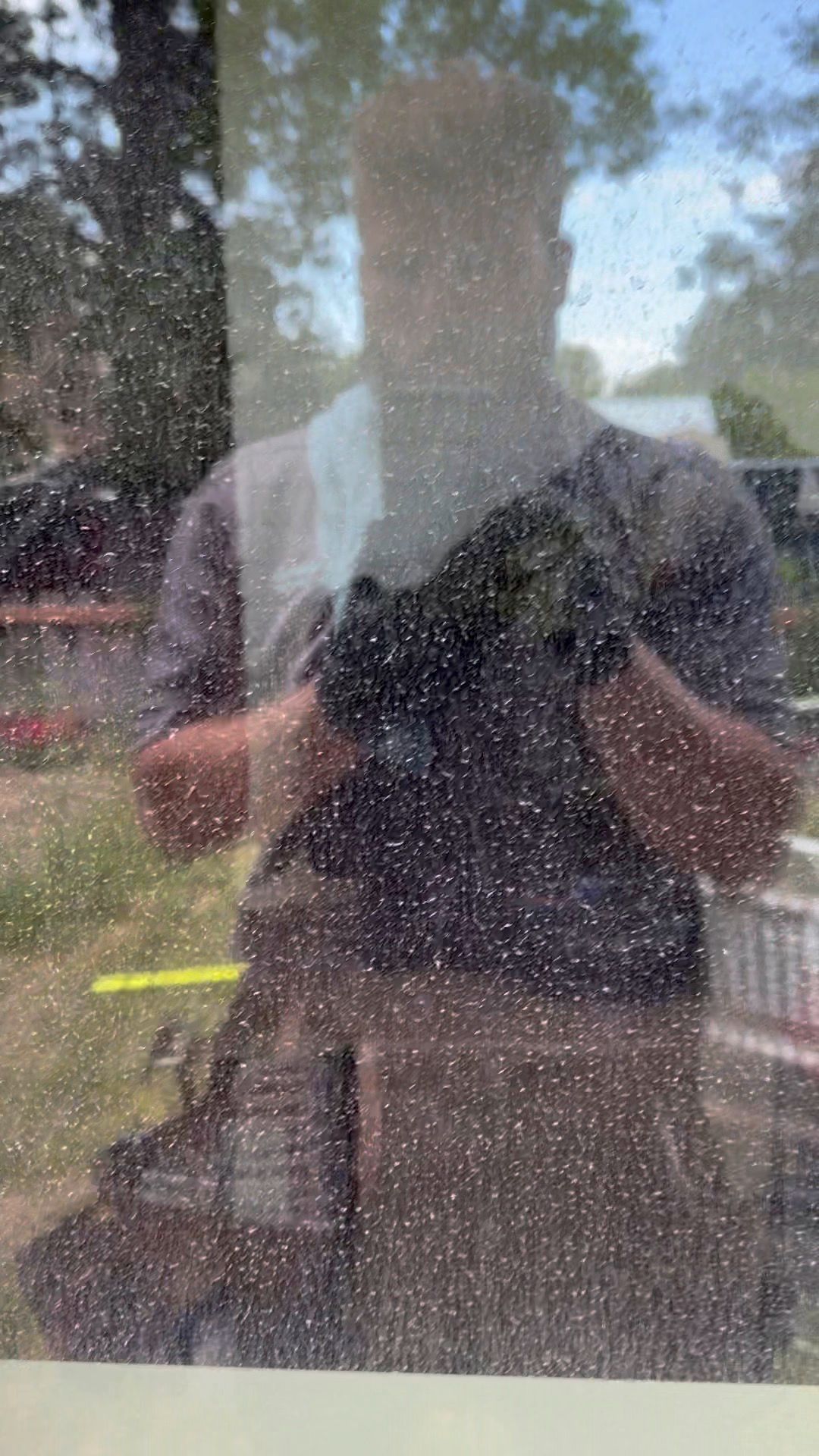Person taking a mirror selfie outdoors, wearing a dark shirt, with a bright sky and trees reflected in the glass.