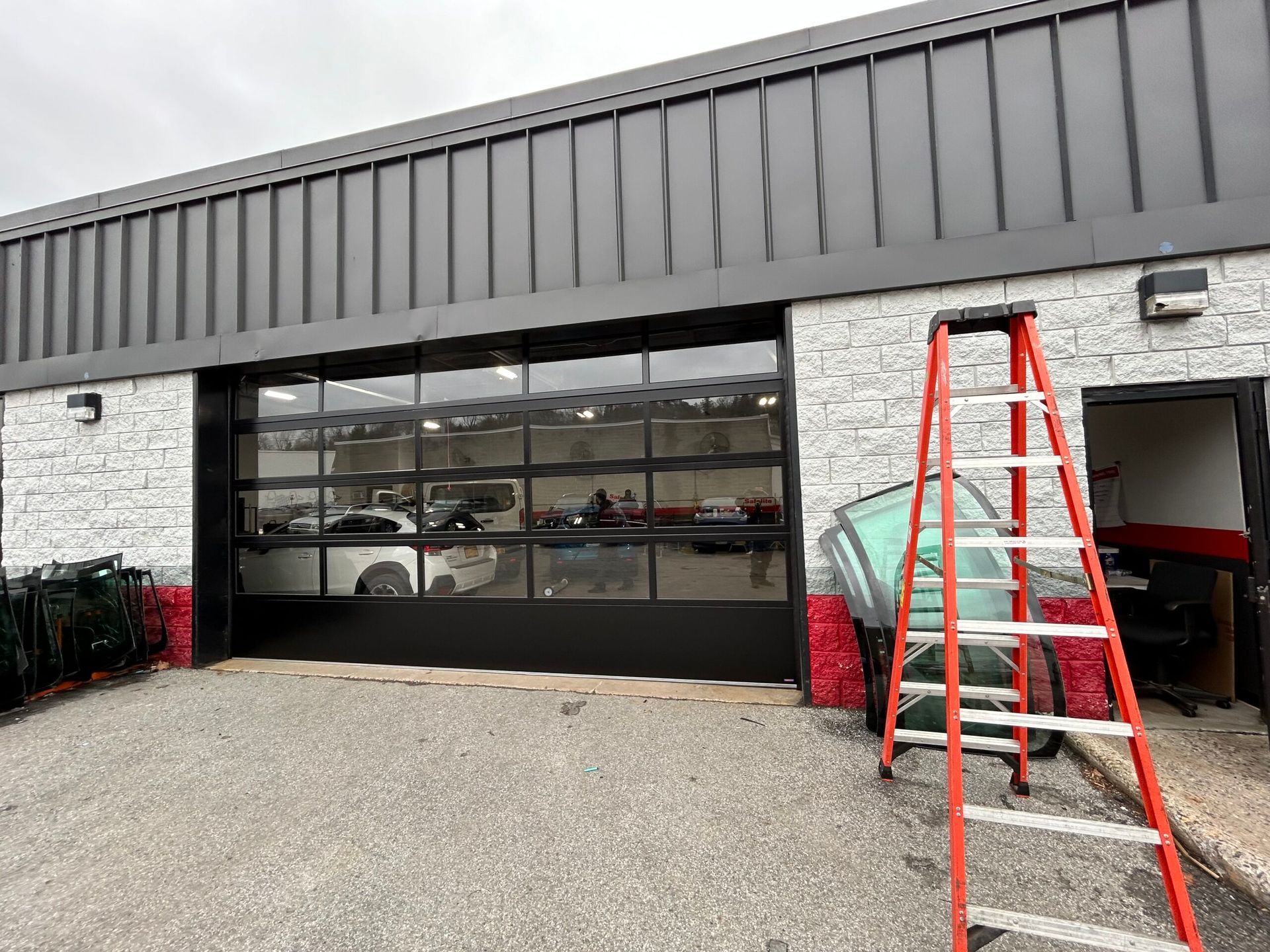 Commercial garage with large glass door, red ladder outside, and parked cars visible inside.