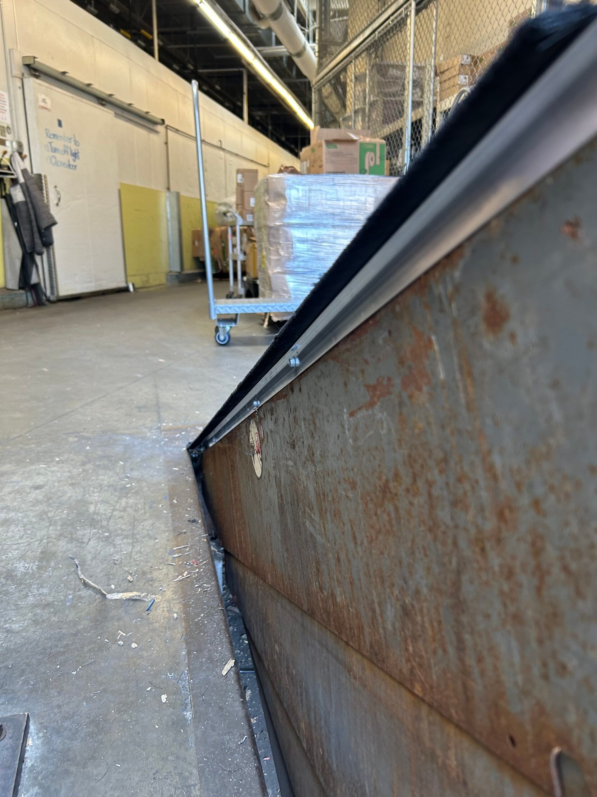 Warehouse aisle with a rusty metal dumpster edge in the foreground and a pallet of wrapped goods in back
