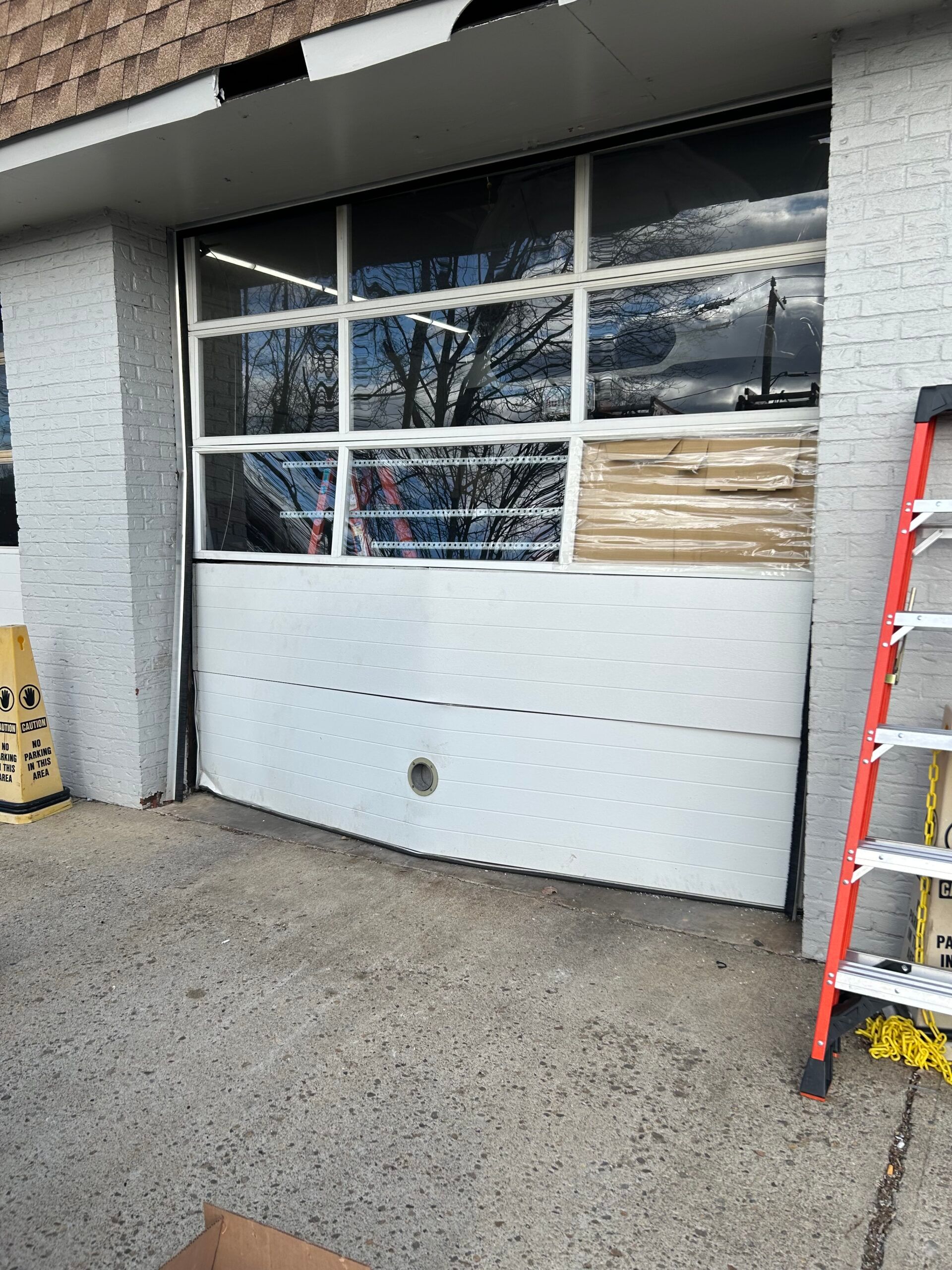 Closed garage door with broken lower panel, reflected windows, and a red ladder on the right.