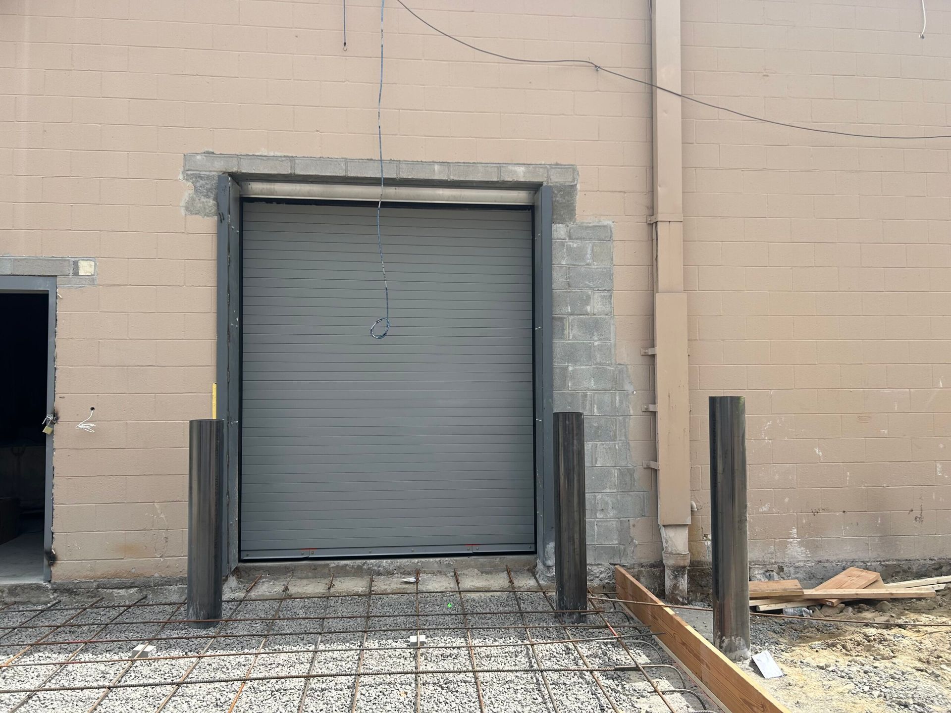 Gray roll-up door set in a beige brick wall, with concrete bollards and an empty loading area.