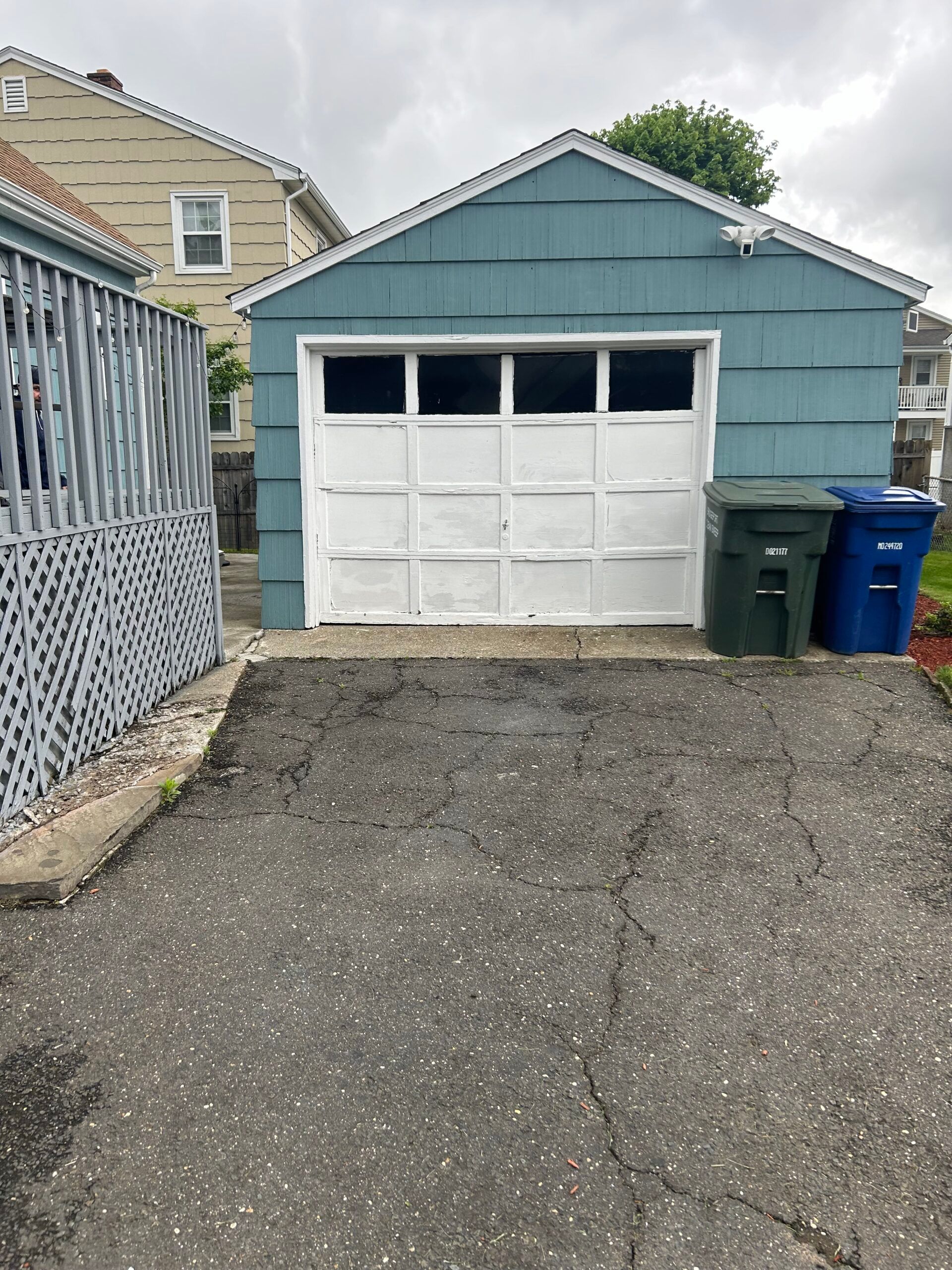 Blue garage with white door and two trash bins beside a gravel driveway and fence.